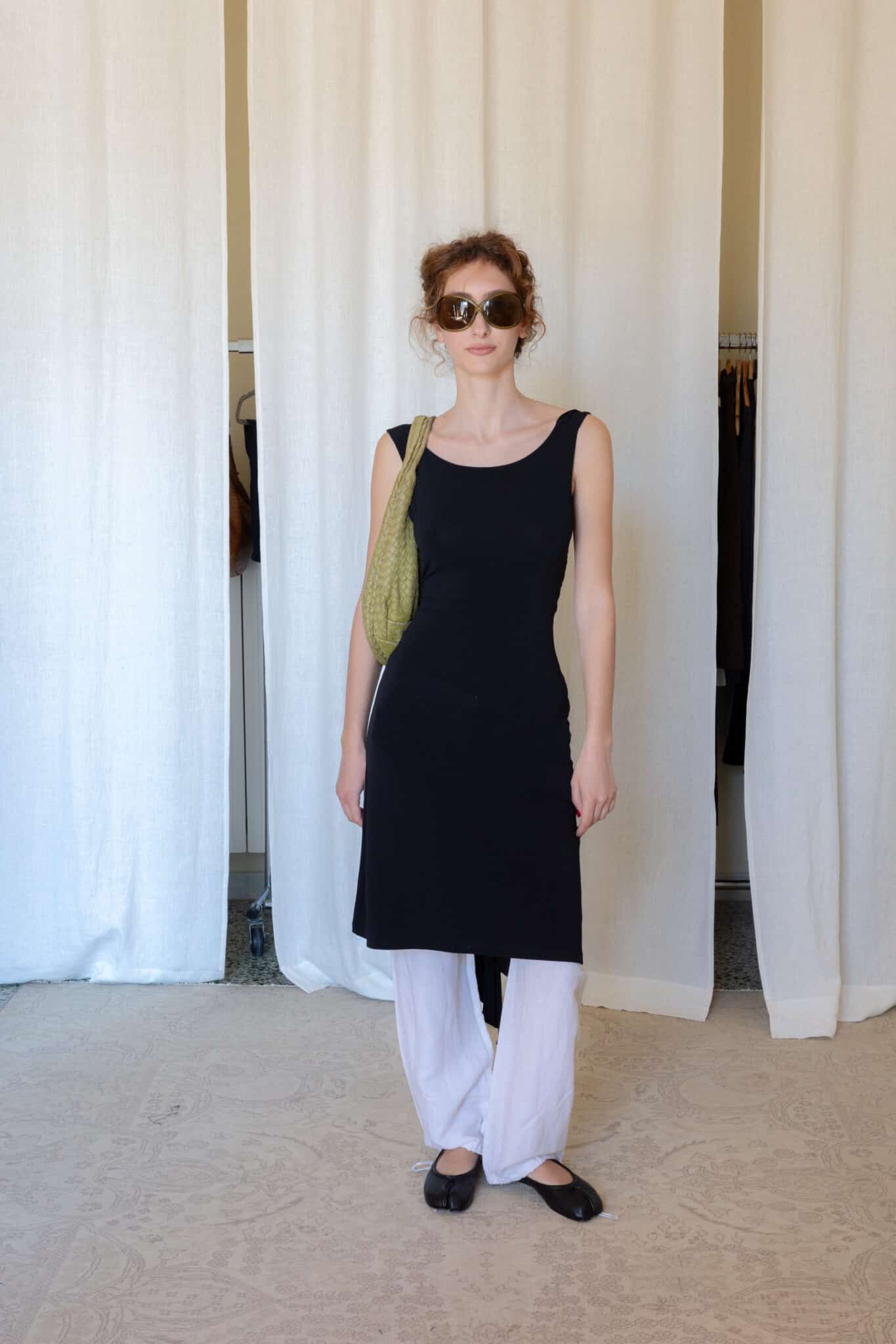 Black open back midi dress layered over white drawstring trousers with black ballet flats and a woven olive shoulder bag.