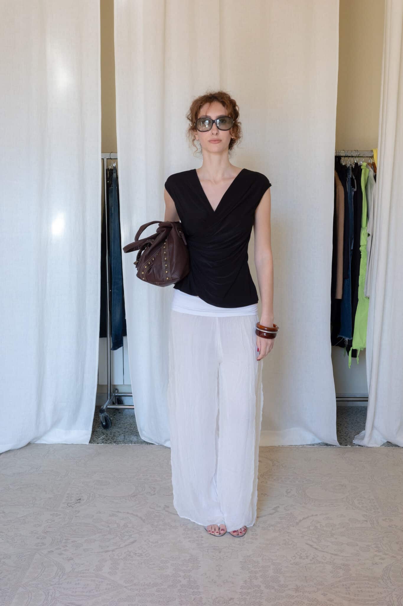 Black draped sleeveless top with a gathered wrap front paired with sheer white wide leg trousers and a dark brown leather bag.