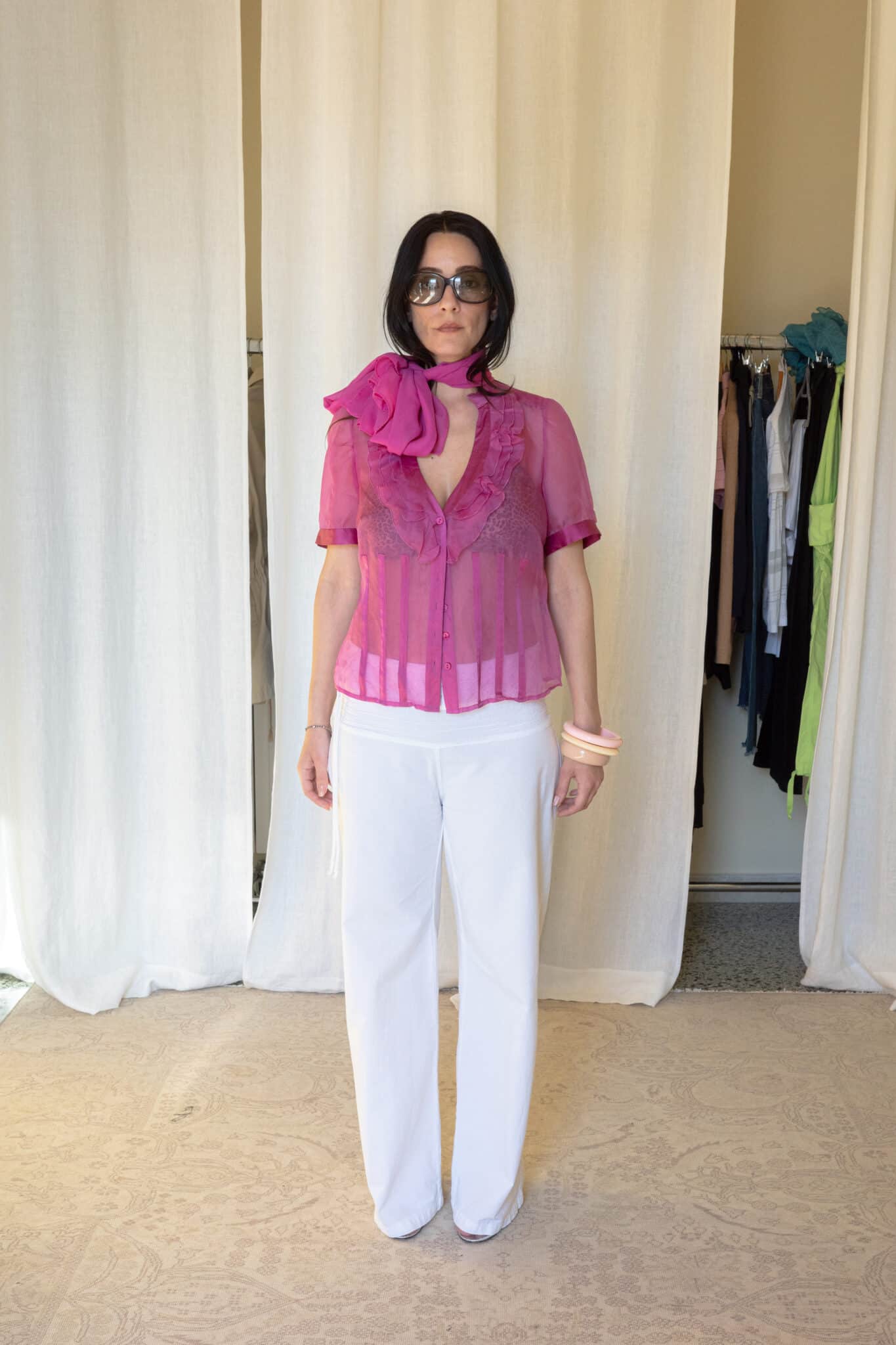 Full outfit featuring a sheer pink silk blouse with layered ruffle detailing along the neckline, matching oversized silk neck scarf and transparent finish, styled with white straight-leg trousers and translucent heels.