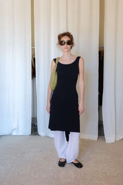Black open back midi dress layered over white drawstring trousers with black ballet flats and a woven olive shoulder bag.