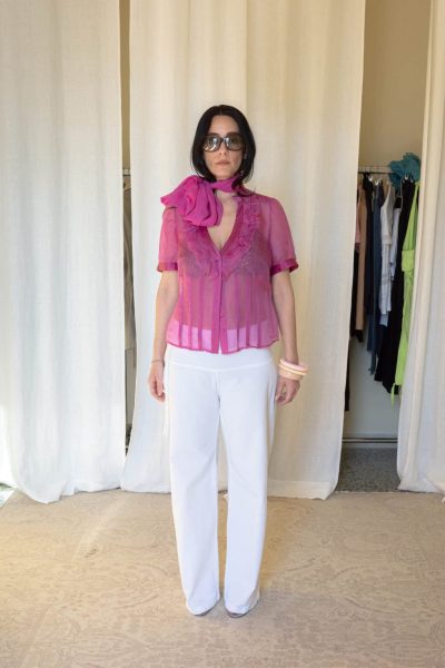 Full outfit featuring a sheer pink silk blouse with layered ruffle detailing along the neckline, matching oversized silk neck scarf and transparent finish, styled with white straight-leg trousers and translucent heels.